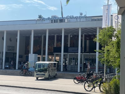 This image depicts the entrance to Palatinus Strand, a thermal bath and outdoor pool complex located on Margaret Island in Budapest, Hungary. The building features a white, modern facade with a colonnade supporting a flat roof. A large sign with blue, stylized lettering reads "Palatinus Strand" above the entrance. Two large white banners with Hungarian text are hanging from poles to the right of the building.
In the foreground, a white electric shuttle vehicle, labeled "PALÁ JÁRAT PALATINUS SHUTTLE SERVICE," is parked on the left. To its right, a woman wearing a blue top and helmet is riding a black bicycle. A rack of bicycles is visible behind the shuttle, with several bikes parked. The ground is paved, and there are some trees and greenery around the area. The sky is blue with scattered clouds.
On the left side of the building, a sign in Hungarian and English indicates "GYÓGYFÜRDŐ ÖLTÖZŐ, MEDENCÉK THERMAL BATH CHANGING ROOM, POOLS." People are seen walking in and around the entrance area. The overall scene suggests a sunny day at a recreational facility in Budapest.