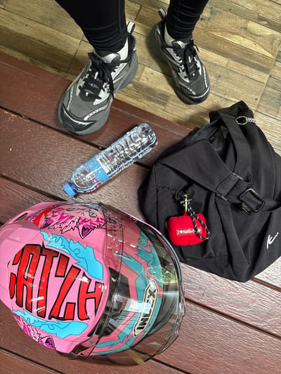 In the city of Ban Kan Chok 23, Thailand, a scene unfolds on a wooden deck. Two feet clad in black leggings and gray and black athletic shoes are visible, one positioned slightly ahead of the other. Nearby, a clear plastic bottle of water lies on its side. To the right, a black duffel bag is placed, adorned with a small red fuzzy pouch bearing the text "I <3 Hello K." A pink motorcycle helmet with a bold red and blue graphic design, featuring the word "CHZZH" in large red letters, rests in the foreground. The environment is an outdoor setting with a wooden floor.