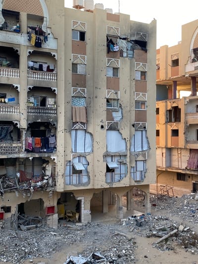 The image depicts a heavily damaged building in Khan Yunis, Palestinian Territories. The building has several floors with extensive structural damage, including holes in the walls and missing sections. Windows are broken or missing, and some openings are covered with white sheeting. The exterior walls are pockmarked with what appear to be bullet holes or shrapnel damage. Debris and rubble are scattered around the base of the building. In the background, another damaged building can be seen. Laundry hangs from balconies on lower floors, suggesting recent habitation.