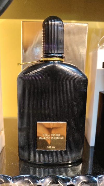 A black Tom Ford "Black Orchid" perfume bottle, 100 ML, stands upright on a dark, reflective surface within Barza Municipality, Syria. The bottle is cylindrical, featuring vertical fluting on its glossy black body and a ribbed, black cylindrical cap. A thin golden cord is tied around the bottle's neck. A gold-colored rectangular label is affixed to the front, displaying "TOM FORD BLACK ORCHID" and "100 ML" in black text. The background is out of focus, revealing a yellow-gold surface on the left and a vertical, light-colored reflective object on the right. No individuals or active interactions are present.