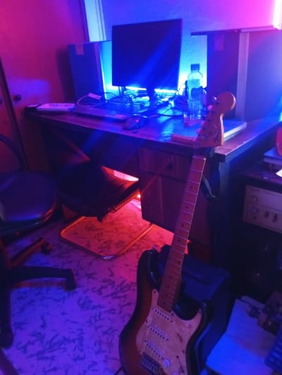 A computer monitor sits atop a cluttered desk in a room illuminated by blue and pink lighting. A laptop, mouse, water bottle, notebook, and speakers share the space. A black rolling chair sits to the side. A sunburst Stratocaster-style electric guitar leans against a black amplifier at the desk's edge. The room appears to be a home studio setup.