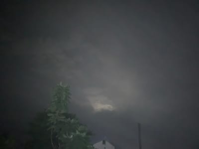 This is a low-light, wide-angle photograph taken at night in Kuru Karama, Nigeria. The sky is filled with dark, heavy clouds, obscuring most of the moon, though a bright, hazy glow emanates from behind the clouds in the upper center of the frame. In the lower left, a portion of a leafy green tree is visible, its branches reaching upwards. In the lower center, the peak of a white building with a dark roof is visible. To the right of the building, a tall, thin, dark vertical pole stands against the cloudy sky. The overall scene is dark and atmospheric, suggesting a cloudy night with limited visibility.