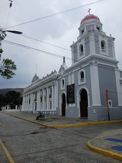 This image shows the Basílica Catedral de Nuestra Señora del Socorro de Valencia in Valencia, Venezuela. The building is a large, ornate structure with a gray and white facade, a red dome, and a prominent bell tower. The sky is overcast. In the foreground, there is a street paved with cobblestones, and a yellow curb. A bench is visible along the building's side. A mountain range is visible in the background. A light pole with wires is on the left side of the frame.