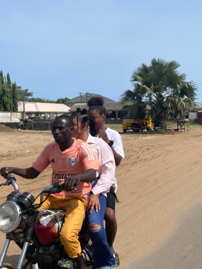 A motorcycle is moving down a dusty road in Ipota, Nigeria. The motorcycle is ridden by a young man wearing a peach-colored shirt and yellow pants. There are two female passengers behind him. One female passenger is wearing a white shirt, the other a pink shirt and jeans with a hole in the knee. A red helmet is on the motorcycle. In the background are buildings and a palm tree.