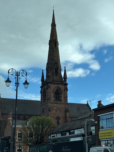 This media file depicts a street view in Chester, United Kingdom, focusing on a prominent church spire. The scene is set against a partly cloudy blue sky, suggesting it's daytime. The spire, constructed from red brick, is a dominant feature, with intricate architectural details visible, including pointed arches and decorative stonework.
In the foreground, a traditional street lamp with two ornate light fixtures stands tall, its dark metalwork contrasting with the sky. Adjacent to the church, modern buildings with brick facades and large windows are visible, some housing businesses. A yellow sign with black text is attached to one of the buildings, reading: "Servicing and repairs book now at Fettle.cc". Another sign, partially obscured, appears to say "Roadside". A tree with sparse spring foliage is situated near the church, adding a touch of nature to the urban landscape. The overall impression is of a historic city center with a blend of old and new architecture. No people are clearly discernible in the image, and no specific event is taking place; it appears to be a general street scene.