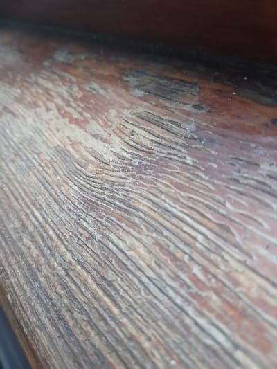 The image shows a close-up, angled view of a weathered wooden surface. The wood grain is prominent, with textured lines running diagonally from the lower left to the upper right. The surface exhibits a reddish-brown base color with lighter, peeling areas and darker, ingrained lines. There are also some lighter specks and a faint web-like structure visible on the surface in the upper right portion of the image. The background is a darker, out-of-focus area, suggesting the edge of a larger wooden object or a shadowed area. There are no people or discernible objects other than the wooden surface itself. The location in Poltava, Ukraine is not visually represented in the image.