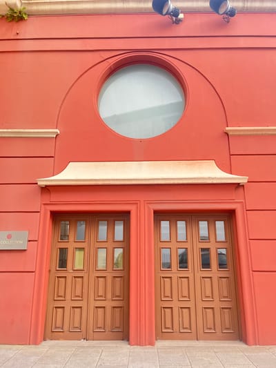 The image shows the exterior of a building in A Coruña, Spain. The building has a bright orange facade and features a large circular window with a light-colored frame. Below the window, there is a decorative architectural element resembling a cornice, also in a light color. Centered below this element are two large wooden double doors, each divided into multiple rectangular panels. To the left of the doors, mounted on the orange wall, is a silver plaque with a logo and the text "COLLECTION". The building's facade is textured and has horizontal decorative lines. Two spotlights are mounted on the upper part of the facade, pointing downwards. The ground in front of the building is paved with light-colored square tiles. A small patch of green vegetation is visible on the top left corner of the facade. The scene appears to be an entrance to a commercial establishment.