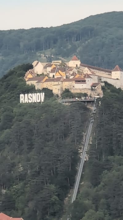 The image shows the Rasnov Citadel in Rasnov, Romania. The citadel is a medieval fortress perched on a hilltop, overlooking a forested valley. A large sign with the word "RASNOV" in white block letters is visible on the slope below the citadel. A funicular railway track ascends the hill towards the citadel, and a viewing platform is located on the side of the hill. The citadel itself consists of several stone buildings with red-tiled roofs and defensive walls. The surrounding landscape is densely covered with green trees. The sky is pale and overcast. There are no people clearly visible in the image.