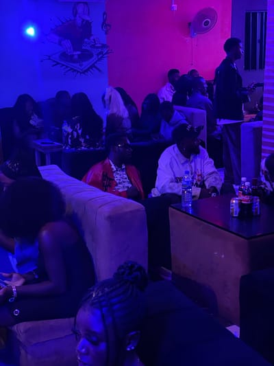 The image depicts a dimly lit, indoor space, likely a lounge or club, with vibrant blue and pink lighting. The walls feature a mural of a DJ and musical notes on the left, and a fan on the upper right. Several individuals are seated on couches and chairs, engaged in conversation or observing their surroundings.
In the foreground, a woman with braided hair is partially visible on the right, looking down. To her left, a person in a bright red jacket with intricate white patterns on the inner lining is seated. In the mid-ground, a man in a white hooded sweatshirt with handprints and text on it is seated, with a bottle of water and cans on the table in front of him. Other individuals are scattered throughout the scene, some partially obscured by the low light and framing of the shot. The overall atmosphere suggests a social gathering. The location is identified as Durumi, Nigeria.