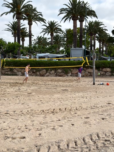 This image depicts a sandy beach scene in Mont-roig del Camp, Spain, under an overcast sky. In the foreground, two males are on a light brown sand beach, marked with numerous footprints and tire tracks. A yellow volleyball net, bearing "COPAYA" branding multiple times, is strung between two wooden posts. The left male, shirtless with white shorts, walks barefoot towards the right, looking down at his right hand. The right male, in a multi-colored t-shirt and grey shorts, is barefoot and captured in motion, appearing to have just kicked a yellow and white volleyball that is visible near the right wooden post. An orange soccer ball and a piece of dark clothing lie on the sand near the right post.
Behind the net, a low wall constructed of light-colored rocks supports a dense, trimmed line of dark green bushes. Above this vegetation, numerous tall palm trees with robust trunks and extensive green fronds dominate the background. Interspersed among the trees and behind the bushes are several recreational vehicles (RVs) and camping structures. A prominent grey RV with a dark rear window is visible to the left of the right volleyball pole, also displaying "COPAYA" branding. A white RV with "KNAUS Sport" text is visible further to the right. A tall metal pole with a security camera or sensor and a white satellite dish extends above the RVs and into the palm trees. The sky is uniformly covered with light grey clouds.