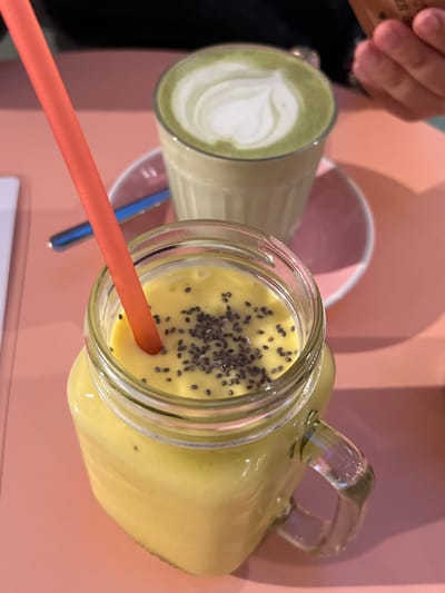 This image displays a close-up, high-angle view of two beverages positioned on a light coral pink table, located in Aldoar, Foz do Douro e Nevogilde, Portugal. In the immediate foreground, a clear glass jar with a handle contains a thick, yellow-orange smoothie generously topped with black chia seeds. An orange plastic straw is inserted into this beverage. Slightly behind and to the right, a clear glass holds a green latte, possibly matcha, adorned with white latte art resembling a heart or stylized leaf pattern. This glass rests on a light pink saucer. A blue spoon is partially visible on the pink table surface between the two drinks. On the upper right side of the frame, the fingers and palm of a person's right hand are visible, holding a small brown packet. The scene suggests an indoor cafe or restaurant setting.