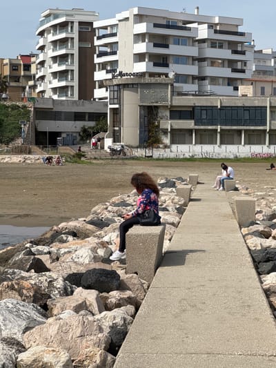 The image captures a scene at Nettuno, Italy, showing a beachfront setting with concrete structures and buildings in the background. The foreground features a woman with curly hair leaning over the edge of a concrete pathway or pier, looking down towards rocks and the water. She's wearing a floral-patterned shirt and black leggings. Another person is sitting further along the path. The background includes modern buildings with white balconies and a mix of architectural styles. The beach is sandy with people relaxing and walking in the distance. The sky is light blue with some clouds.