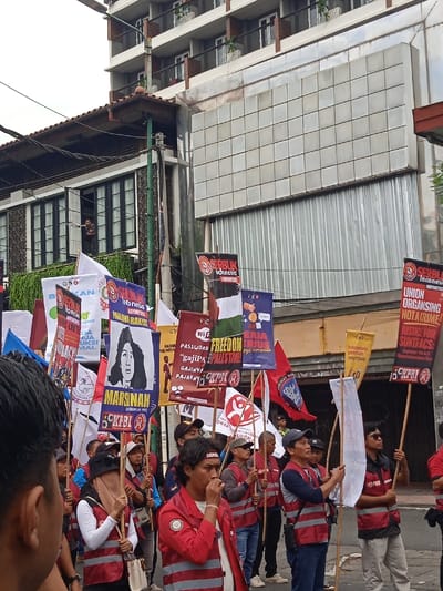 The image depicts a protest taking place in front of a building located at latitude -7.784 and longitude 110.367 in the city of Yogyakarta, Indonesia. The building appears to be a multi-story, modern structure with a tiled facade. The protesters, mostly men, are holding aloft various signs and banners with slogans written in Indonesian. Some of the slogans are visible and include "Freedom Palestine", "Password 'Gajipa' & All the Time", "Union Organising Not A Crime!", and "Justice for Sunticas". The protesters are wearing red vests and carrying wooden poles with flags and signs attached to them. They appear to be standing on the street in front of the building, waiting for instructions or marching in a procession. The atmosphere is one of anticipation and determination. The signs and banners are clearly visible, as are the protesters’ faces, which express a mixture of resolve and excitement. There is a sense of collective action and shared purpose.