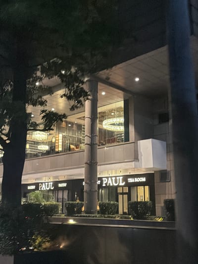 The image shows the exterior of a PAUL bakery and tea room located in Gurgaon, India. The establishment features illuminated signage with the "PAUL" logo, accompanied by "BAKERY" and "PASTRY" on one side, and "TEA ROOM" on the other. The building has large glass windows, showcasing elegant interior lighting fixtures, including large circular chandeliers. The exterior is lined with well-maintained green bushes and some palm trees. The scene is captured at night, with artificial lighting highlighting the storefront. A large concrete pillar is prominently visible in the foreground. The overall impression is of a well-lit, modern establishment in an urban setting.