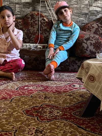 Two young children are visible in the image. One child, wearing a light blue pajama set with orange cuffs and a pink hat, is seated on a floral-patterned sofa with their feet extending onto a patterned rug. A multi-socket power strip is situated near the sofa. The second child, wearing light-colored pajamas with pink patterned pants, is lying down. A brick-patterned wall is in the background. A table partially covered with a patterned tablecloth is nearby. The location is in a domestic interior, likely in Khan Yunis, Palestinian Territories.