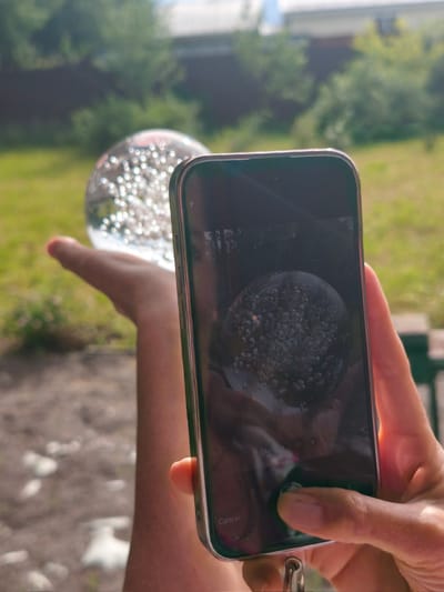 This media file shows a close-up of a person's hand holding a crystal ball filled with small bubbles. The crystal ball is transparent and refracts light, creating a shimmering effect. In the foreground, a smartphone is held in the other hand, with its screen reflecting the crystal ball and its surroundings. The phone screen displays a captured image of the crystal ball, showing the bubbles magnified.
The background is out of focus, but suggests an outdoor setting with greenery, possibly a garden or yard, with hints of a building or fence. The lighting indicates it is daytime, likely sunny given the bright reflections. The presence of a phone being used to capture an image of the crystal ball suggests the activity of photography or videography, perhaps for artistic or observational purposes.
The image evokes a sense of curiosity and perhaps wonder, as the viewer is presented with a close-up perspective of the crystal ball and its intricate details. The reflection on the phone screen adds a meta-layer, showing the act of capturing the image itself. There is no visible text other than the word "Cancel" which appears on the smartphone screen, likely part of the camera interface. The location context of Steklyannyi, Russia, places the scene within a specific geographical area, though no direct visual cues confirm this.