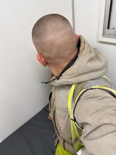 An overhead-angle image depicts an individual from behind, located within an indoor setting in Eastleigh, United Kingdom. The subject exhibits a very short, buzz-cut hairstyle with visible scalp and ears. They are dressed in a light beige hooded jacket, which appears soiled, with a black drawstring visible near the neck. Over this jacket, a bright yellow high-visibility vest featuring grey reflective stripes is worn. The vest appears secured with black fasteners. A dark-colored garment is visible underneath the beige jacket's collar. The immediate environment consists of plain white walls. A dark grey surface, possibly a bench or floor mat, is visible in the lower left. In the upper right background, a partially obscured metallic-framed window or opening is present. No discernible actions or interactions are observed.