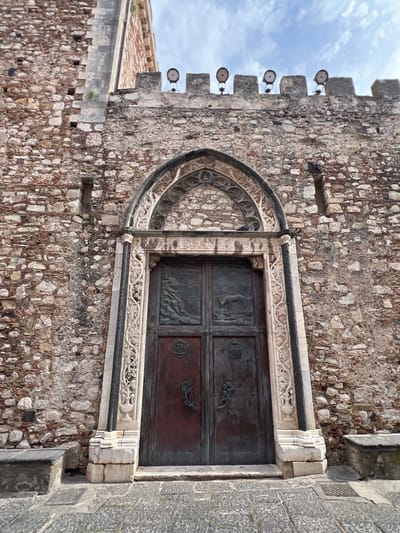 An eye-level, medium shot captures the ornate, dark-colored double doors of a stone building in Taormina, Italy. The doors are set within a grand stone archway featuring intricate carvings. Above the arch, the stone wall of the building rises, topped by a crenellated parapet with several decorative light fixtures. The wall itself is constructed of rough-hewn stones of varying sizes and colors, with visible mortar. To the left of the doors, a rough stone bench is visible. The ground in front of the building is paved with cobblestones. The sky above is partly cloudy.
