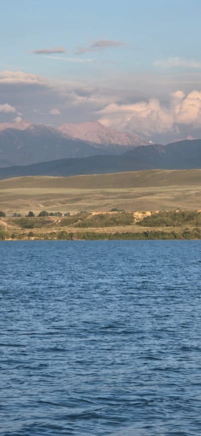 The image depicts a serene natural landscape. The foreground is dominated by a vast expanse of blue water, with gentle ripples disturbing its surface. Beyond the water, a treeline borders the shore, giving way to rolling hills covered in dry, golden-brown grass. Further in the distance, layered mountain ranges rise, their peaks softly illuminated by the sky. The sky itself is a pale blue, with scattered clouds, some appearing wispy and thin, others more substantial and tinged with pink. The overall impression is one of tranquility and natural beauty.