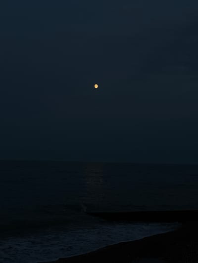 The image depicts a nighttime scene at Felixstowe, United Kingdom. The primary subjects are the dark, expansive sky with a faint, yellowish moon positioned to the left, and the dark, textured surface of the sea on the right, with a visible wave cresting and breaking. The scene is very dark, suggesting it is either late night or early morning. The weather appears to be clear, as the moon is visible and no clouds obscure the sky.
The setting is clearly a coastal location, indicated by the presence of the sea and waves. The overall mood is serene and perhaps a little melancholic due to the darkness and the solitary moon. The rough texture of the water and the faint shimmer on its surface suggest movement and depth. There is no visible text in the image.