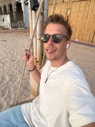A young Caucasian male with blond, spiked hair and sunglasses is sitting on a swing on a sandy beach. He is wearing a white t-shirt, blue jeans, and white earbuds. He has his tongue sticking out and is holding onto the rope of the swing with his left hand. Behind him, there is a bamboo wall and a building under construction. The image was taken in Choeng Thale, Thailand.