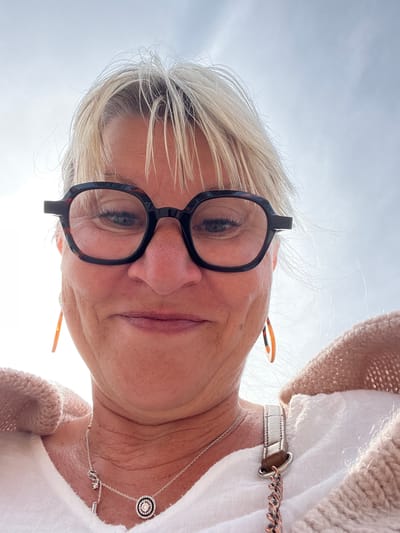A close-up, low-angle shot of a blonde woman wearing black-rimmed glasses and a white shirt with a beige sweater. She is also wearing a silver necklace. The photo seems to be taken outdoors under a bright sky, possibly Palavas-les-Flots, France, given the prompt's mention. There are no actions or interactions depicted other than her posing for the photo.