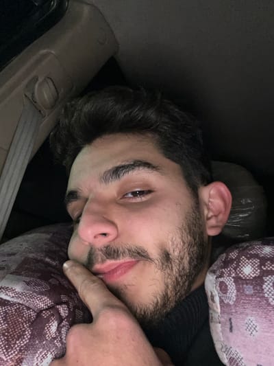 A man with dark hair and a beard reclines against patterned pillows and possibly the interior of a car. He is turned to face the camera with one eye open. His right index finger touches his chin or cheek. A tan interior panel of a vehicle is seen near his head and a dark plastic bag or crumpled material rests behind him. The lighting is low.