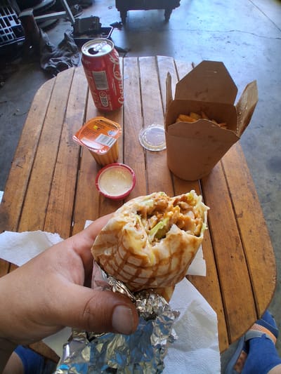 A first-person perspective shot shows a person holding a half-eaten shawarma. The shawarma is wrapped in a white tortilla and filled with meat, lettuce, and sauce. It's partially unwrapped and resting on aluminum foil and a white napkin.

On a wooden table, to the left of the shawarma, is a small red container with a creamy sauce. Further left, there's an orange-lidded plastic cup, possibly containing sauce or dressing, with a label that includes a barcode. To the right of the shawarma, a cardboard takeout box is open, revealing a portion of french fries. Behind that, an open red Coca-Cola can is visible.

The table is made of wooden planks. In the background, a concrete floor is visible with some cracks and debris. The overall setting appears to be outdoors, possibly a casual dining area or a rest stop. The image does not contain any visual elements that definitively place it in Mas Aliu, Spain, or any other specific city.