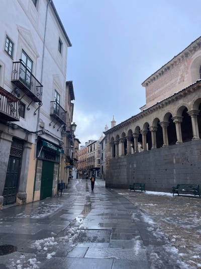 The image shows a street in Segovia, Spain (latitude: 40.949, longitude: -4.122). The street is narrow and cobbled, with a white building on the left and a stone building on the right. The stone building features a row of arches. The street is wet and there is snow on the ground. A single person, dressed in black, walks down the street. Two benches are visible, one on the left and one on the right. There is a sign on the left building reading "Salchicheria Briz." The sky is overcast.