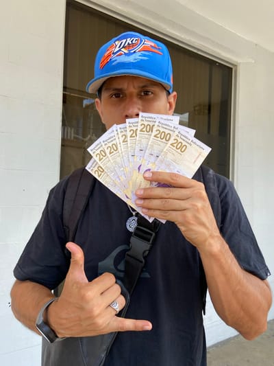 A man stands in an outdoor setting, possibly in San José de Barlovento, Venezuela, holding a fan of Venezuelan Bolivar notes in front of his face. The notes are 200 Bolivar denominations. He wears a blue baseball cap with an orange logo, a black t-shirt, and a black shoulder bag. He is making a hand gesture. He also has a ring on his finger.