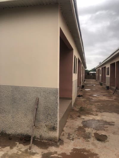 The image shows a view down a narrow walkway between two identical, single-story buildings with light beige and pink facades and corrugated metal roofs. The walkway is paved with concrete that is wet and stained, with several puddles visible. Black sandals are scattered on the ground, and two green buckets are placed near the entrance of one of the buildings. A rough, textured grey material covers the lower portion of the building walls. In the foreground, leaning against the building on the left, is a vertical wooden pole with bamboo-like segments. The sky is overcast and grey. The buildings appear to be part of a row of housing units or possibly classrooms.