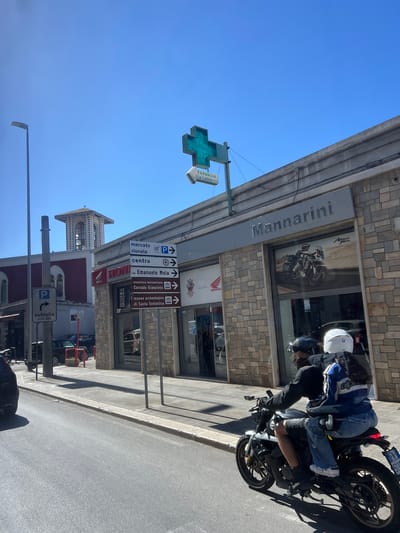 The image captures a vibrant street scene on a clear, sunny day, featuring a building with a stone facade, a person on a motorcycle, and various street signs. The sky is bright blue and cloudless.
The primary subject is a person seated on a black and grey motorcycle, wearing a white helmet, a blue and black protective jacket, jeans, and gloves. Another person or passenger is partially visible behind them. The motorcycle has a visible license plate with "511" clearly legible.
The building on the left houses a "Mannarini" dealership, indicated by a prominent sign and a "HONDA" logo. A poster for "Africa Twin" motorcycles is visible in the window. A green cross pharmacy sign, displaying "FARMACIA Via Lattanzio," hangs prominently.
Several directional and informational signs are mounted on poles. These signs indicate local points of interest and services, including "P mercato rionale" (market parking), "P centro" (city center parking), "via Emanuele Mola," "pinacoteca metropolitana Corrado Giaquinto" (art gallery), and "museo archeologico di Santa Scolastica" (archaeological museum). Another sign indicates "P autosilo a m. 250" (parking garage 250 meters away). A bell tower is visible in the background, adding to the architectural context. The building itself has the number "146" on its facade. The bright light suggests it is midday or early afternoon. The signs are in Italian, confirming the location in Italy, specifically Bari.
