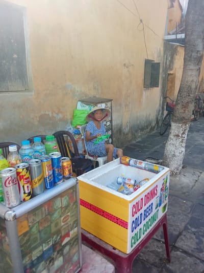 In Hội An Ward, Vietnam, an elderly woman wearing a conical hat and a blue and white checkered dress sits on a plastic stool next to a cooler full of drinks. The cooler is yellow and has "COLD DRINKS", "COLD BEER", and "COLD COCONUT" written on its side. Various cans and bottles of beverages are visible on a display stand in front of the woman, including Coca-Cola, Schweppes, and Tiger beer. In the background, a tree trunk with white bark is prominent, and bicycles are parked nearby. The scene appears to be a street-side vendor stall.