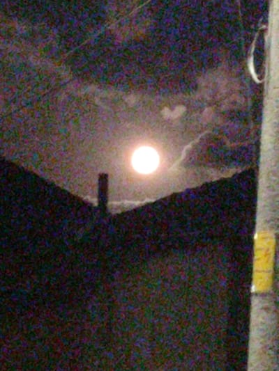The image depicts a nocturnal scene dominated by a bright, circular moon against a dark, cloudy sky. In the foreground, a silhouetted structure, possibly a tower or chimney, rises from a dark, hilly landscape. To the right of the frame, a pole with yellow markings is visible. The overall image quality is low, with significant digital noise and a lack of sharp detail, suggesting it was taken in low light conditions. The specific location in Smolyan, Bulgaria, cannot be determined from the visual information provided.