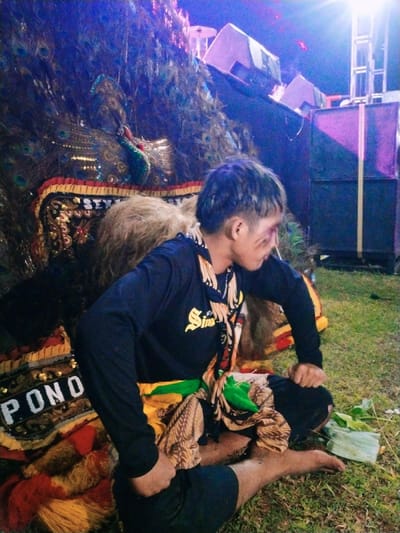 A young man sits on the ground, his legs crossed. He is wearing a black long-sleeved shirt with a gold design on the front, and a yellow and green sarong. He is holding his left hand in the front of his pants and his right hand is resting on his lap. The man is seated in front of a large, colorful, feathered display. The display is made of many feathers and has gold and red accents. The scene appears to be set in an outdoor location in the evening. Behind the man are large speakers, a large, purple object and a metal scaffolding tower. The location is in Kecamatan Sambirejo, Indonesia, with coordinates -7.5 latitude and 111.087 longitude. The grass around the man is green and appears to be well-maintained.  The man is sitting in front of a large, colorful, feathered display. The display is made of many feathers and has gold and red accents. It appears to be a traditional Indonesian display.  The scene appears to be set in an outdoor location in the evening. There is a large, purple object and a metal scaffolding tower behind the man. The scene is brightly lit and there appears to be a festive atmosphere.