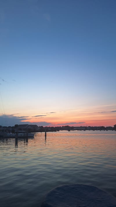 The image depicts a serene sunset over the harbor of Nantucket, Massachusetts, USA. The sky transitions from a deep blue at the top to soft hues of pink and orange near the horizon, with thin, wispy clouds scattered across. The water in the foreground is calm, reflecting the colors of the sky. Several boats, including sailboats and smaller vessels, are moored in the harbor. Buildings and trees line the distant shore, suggesting a coastal town. Two wooden posts rise from the water in the mid-ground, marking a dock or mooring area. The overall scene evokes a peaceful evening atmosphere.
