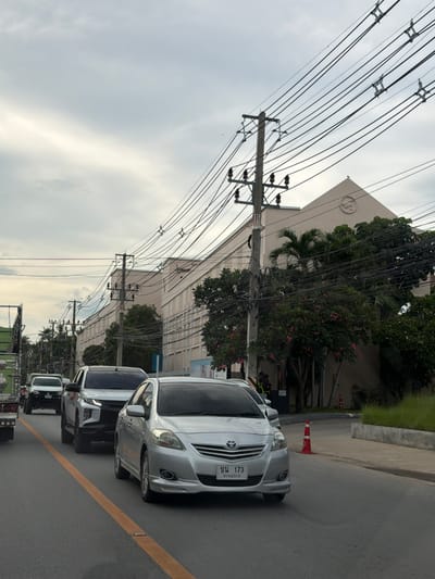 The image captures a street scene in Baan Bo Phut, Thailand, likely during the day, given the ambient light and overcast sky. The foreground features a road with a yellow line marking the lane. Several vehicles are visible, including a silver sedan prominently positioned, displaying a license plate with Thai characters. Behind it, a dark-colored SUV and other vehicles follow, suggesting traffic.
The background is dominated by a long, light-colored building, possibly a commercial or industrial establishment, with trees and palm trees lining its facade. A complex network of overhead power lines stretches across the scene, supported by utility poles, a common sight in many urban and suburban areas. Red and white traffic cones are present on the side of the road, hinting at ongoing work or a temporary traffic diversion. The overall atmosphere appears calm, with no obvious signs of hurried activity. The weather is cloudy, casting a diffused light over the scene.
