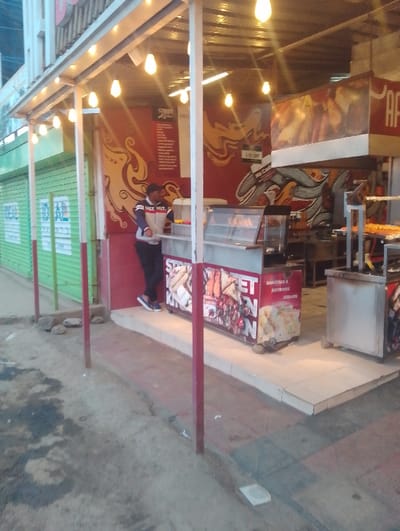 The image shows the exterior of a street food stall in Rungiri, Kenya. The stall is made of white metal and has a red and white awning. The stall is decorated with graffiti-style artwork. A man in a blue and white jacket is standing in front of the stall. He appears to be looking at something off camera. There are several food items visible, including sandwiches and hot dogs, all of which are arranged in a display case that is sitting on a counter. Several items of food, including sandwiches, hot dogs and other fast food are visible. The counter is located under a sign that says "Street Kitchen" and is decorated with the same graffiti-style artwork as the awning. The stall is located on a street with a dirt sidewalk. There are several pieces of trash on the sidewalk. The street is empty except for the man.