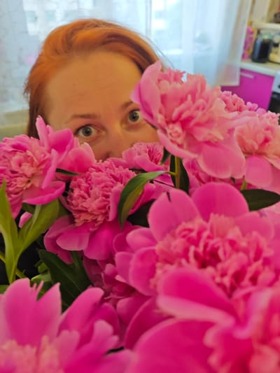 A woman with reddish-brown hair peeks out from behind a large, vibrant bouquet of pink peonies, shot in an indoor setting in Markovskii, Russia. The majority of the frame is filled with the lush, abundant petals of the peonies, with some flowers in sharp focus in the foreground and midground, creating a sense of depth.
The woman's forehead and wide, alert eyes are visible above the flowers, looking directly at the viewer with an expression that could be interpreted as surprised, playful, or curious. Her face is positioned centrally in the upper half of the image, emerging from the dense floral arrangement.
The background is softly blurred, indicating an indoor environment with natural light streaming in from what appears to be a window. On the right side of the background, a hint of bright pink cabinetry or furniture can be seen, adding a splash of complementary color to the scene. The overall lighting is soft and natural, illuminating the woman's face and the rich hues of the flowers. There is no visible text in the image.