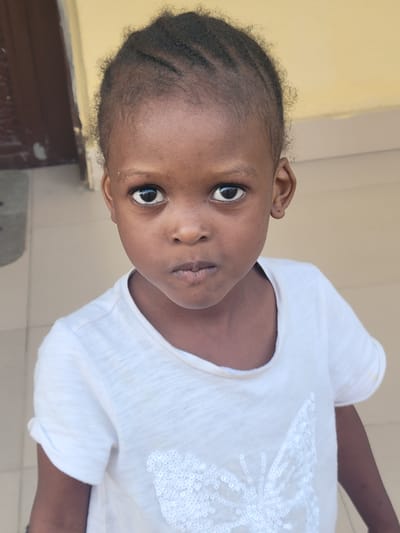 A young Black child with braided hair, wearing a white t-shirt with sequin detail, is looking directly at the camera. The location is Suleja, Nigeria, as indicated by context. The background consists of a light yellow wall and a tiled floor with a dark brown wooden object partially visible in the lower left corner. The child appears to be indoors.