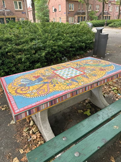The image depicts a mosaic-tiled bench in an outdoor urban setting. The bench features a colorful mosaic design with intricate patterns, including what appears to be a chessboard in the center, surrounded by floral and bird motifs. The bench itself is made of concrete with decorative mosaic tiles along its edges. In the foreground, a green wooden bench is partially visible. The background shows residential buildings with brick facades, trees, and a dense green hedge. A dark-colored trash can is also present. The ground is paved with asphalt and scattered with dry leaves, indicating an outdoor public space, possibly a park or courtyard. The buildings appear to be in the city of Amsterdam, Netherlands, suggested by the architectural style. There are no people visible in the scene.