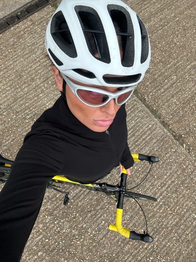 A cyclist wearing a white helmet and pink tinted sunglasses is positioned on a road or path, likely in the city of Tonbridge and Malling, United Kingdom. The person is wearing a long-sleeved black top and holding the handlebars of a bicycle, which is yellow and black. The surface beneath them appears to be a concrete or paved pathway. The view is taken from above, looking down at the subject.