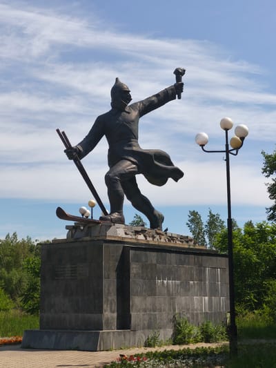 A bronze statue of a skier stands on a large, multi-tiered granite pedestal in Komsomolsk-on-Amur, Russia. The skier, depicted in mid-stride, holds ski poles in their left hand and a torch in their outstretched right hand. The statue is positioned in an outdoor setting with a blue sky dotted with white clouds above. To the right of the statue is a black lamppost with four spherical white lights. The pedestal is inscribed with text in Russian. The surrounding environment includes lush green trees and vegetation, and a paved walkway with some flower beds in the foreground. The overall scene is a monument commemorating an event or figure, set against a natural backdrop under a bright sky.
