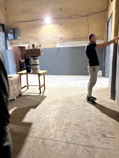 The image captures an indoor scene in what appears to be a construction or renovation area, possibly located in Dupnitsa, Bulgaria. In the foreground, a person is partially visible from the left side of the frame, their dark clothing and shadow dominating the lower left quadrant. The central and right portions of the image focus on a man standing on the right, facing towards a wall and seemingly engaged in an activity involving measuring or marking it. He is wearing a dark t-shirt, light-colored pants, and black and white athletic shoes. His right arm is extended towards the wall.
Behind the man, a wooden table stands in the mid-ground, topped with two stacked white paint buckets and a cardboard box. To the left of the table, a pallet wrapped in plastic film is visible, containing what appear to be stacks of materials. The environment's walls are textured and painted in a combination of light yellow and grey. Overhead, electrical wires and a light fixture are visible, suggesting unfinished electrical work. The floor is a concrete surface with visible seams and some debris. The overall atmosphere is utilitarian, indicative of an ongoing project.