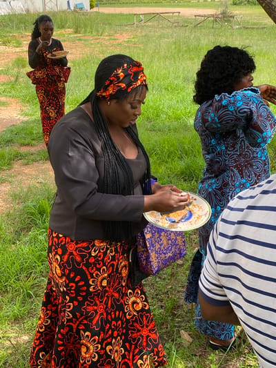 In Kuru Karama, Nigeria, an outdoor gathering is underway on a grassy area with sparse dirt paths and faint wooden structures in the background. Three women are visible. The woman in the foreground, wearing a black top and a vibrant orange and black floral print skirt, is holding a plate of food and appears to be eating. She has long, braided dark hair, accessorized with a matching orange and black headscarf. To her right, another woman, partially visible, is wearing a blue and brown floral patterned dress. Further in the background, a third woman, dressed in a similar orange and black floral print attire as the foreground woman, stands holding a plate. A person wearing a blue and white striped shirt is also present, partially obscuring the view of the woman in the blue dress.