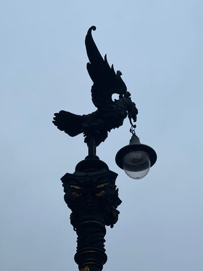 A black silhouette of an ornate lamppost is visible against a pale, overcast sky. The lamppost features a detailed, sculpted top in the shape of a mythical creature, possibly a dragon or a griffin, with wings outstretched and a lamp hanging from its beak. The base of the lamppost is also elaborately decorated with sculpted elements and gold accents. The lamppost is positioned in the city of Bilbao, Spain, likely a decorative feature in a public space. The scene is devoid of any people or discernible activity, focusing solely on the architectural element and the atmospheric sky.