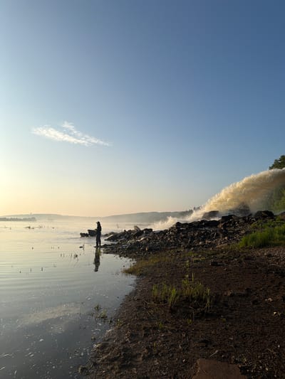 An early morning scene unfolds by a body of water, likely the Ufa River, near Chernushka, Russia. The sky is a clear, pale blue with a wispy white cloud near the horizon. The sun is low, casting a warm, golden glow that softens the landscape.
In the foreground, the dark, damp earth of the riverbank is dotted with sparse green grass. The water's edge is rocky, with large, dark stones creating a natural barrier. A solitary figure, silhouetted against the light, stands on these rocks, fishing with a rod. They are dressed in dark clothing, including waders and a hat. Their reflection is visible in the calm water, which is disturbed by gentle ripples and small patches of foam.
Further out on the water, several small dark shapes, possibly birds or debris, are scattered. The distant shoreline is hazy with mist, obscuring the details of hills and trees.
To the right of the image, a powerful torrent of water is being discharged, creating a dramatic cascade over the rocks. This creates a frothy, tan-colored spray that contrasts with the otherwise tranquil scene. The force of this discharge suggests an industrial or infrastructural process. The overall impression is one of natural beauty punctuated by human activity.
