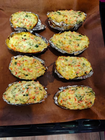 The image shows a baking sheet with eight baked potatoes. Each potato is halved and stuffed with a mixture of potato flesh, herbs, and some red ingredient. The potatoes are nestled in aluminum foil cups. The baking sheet is dark brown and appears to be lined with parchment paper. The image appears to be taken indoors. It is not possible to determine the location in Markovskii, Russia, based on the given image.