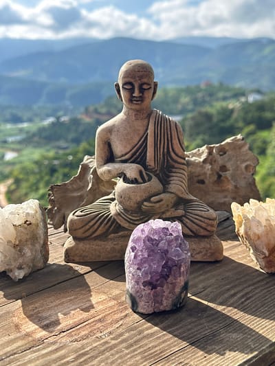 A close-up, eye-level, outdoor shot captures a Buddha statue on a wooden surface with a blurred mountainous landscape in the background. The Buddha statue is positioned centrally, made of a light brown, stone-like material, with its eyes closed and holding a rounded bowl. To the left of the statue, a large, rough, clear crystal cluster is visible, and to the right, a cluster of pale yellow crystals. In front of the statue, a vibrant purple amethyst geode sits prominently. The wooden surface is weathered with visible grain and cracks. The background shows rolling hills covered in lush greenery and a cloudy sky. The scene is set in Thôn 3, Vietnam.