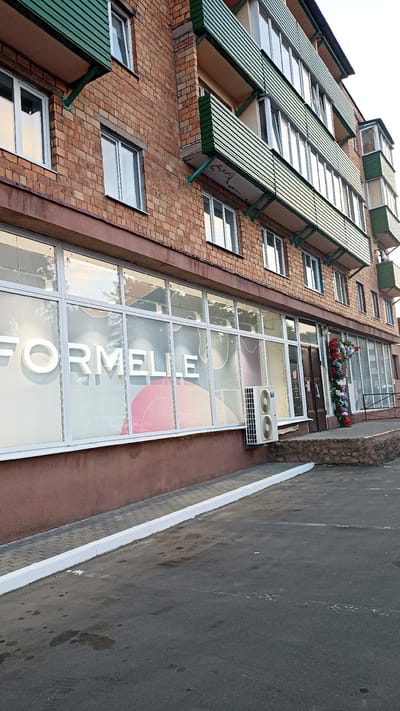 The image shows the exterior of a multi-story brick building in the city of Kapyĺ, Belarus. The building has several floors with multiple windows and balconies with green metal slats. The ground floor features large storefront windows with the text "FORMELLE" visible on one of them. An air conditioning unit is mounted on the exterior wall near the storefront. To the right of the storefront, there is an entrance with an archway decorated with colorful artificial flowers. The ground in front of the building is paved with asphalt and a sidewalk with a white curb. The scene appears to be during daylight hours, with soft lighting.